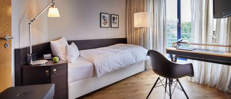 A hotel room with a bed, bedside table, lamp, desk with chair, and a TV.