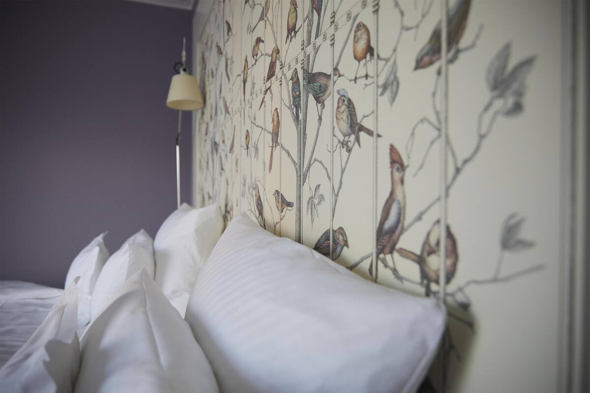 Una cama con ropa blanca y una pared con estampado de pájaros.