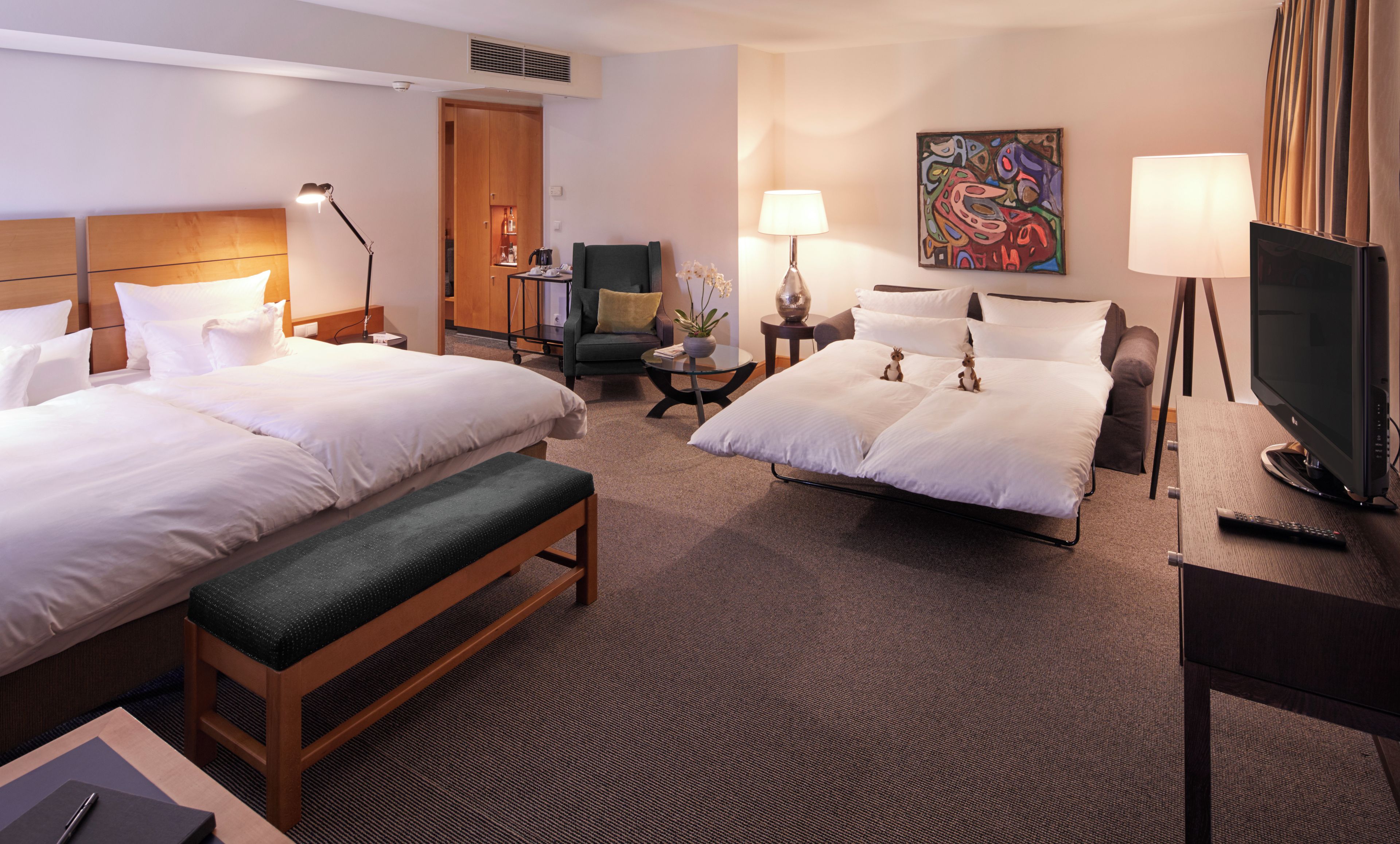 Ein Hotelzimmer mit zwei Betten, einem Schreibtisch, einem Sessel, einem Fernseher und einem Bild an der Wand.