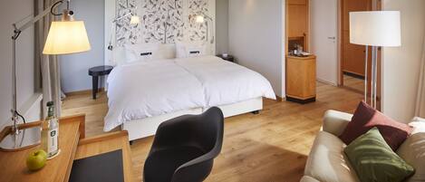 Habitación de hotel moderna con una cama grande, un escritorio con lámpara, una silla y un sofá.