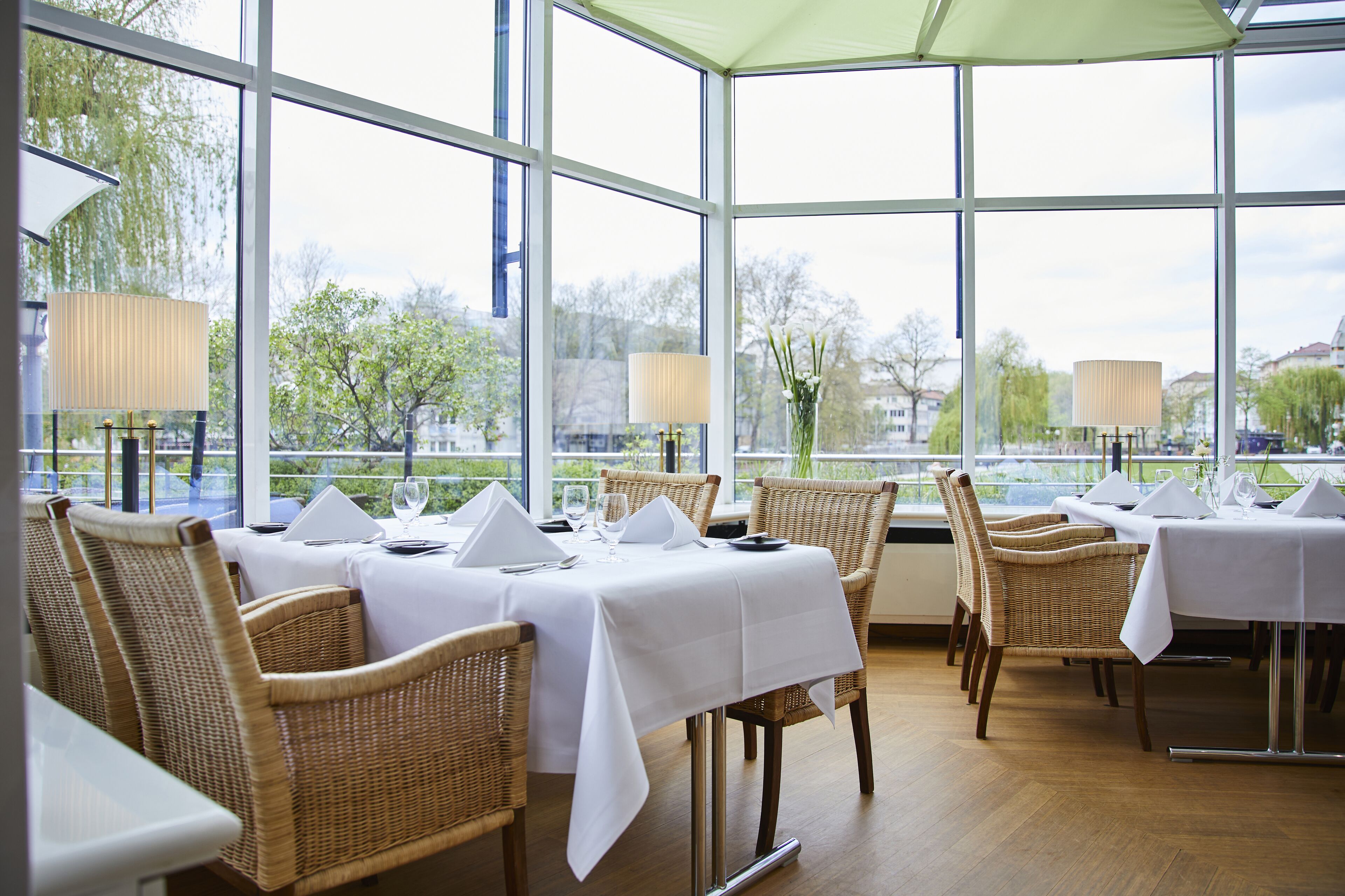 En restaurant med dækketøjborde, fletstole og store vinduer med udsigt til træer og bygninger.