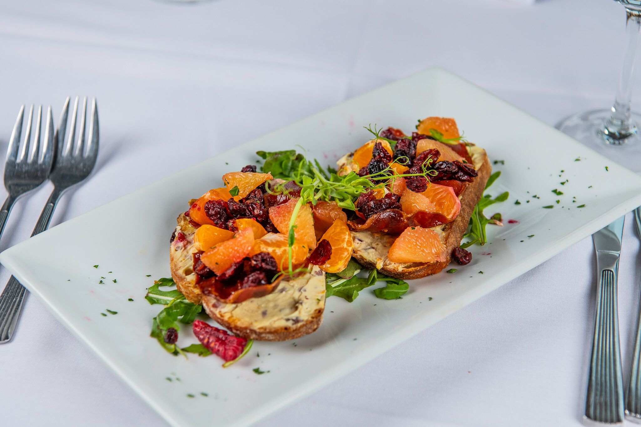 Plato rectangular blanco con un alimento cubierto de segmentos de naranja, arándanos deshidratados y hojas verdes, acompañado de cubiertos sobre un mantel blanco.