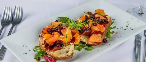 Plato rectangular blanco con un alimento cubierto de segmentos de naranja, arándanos deshidratados y hojas verdes, acompañado de cubiertos sobre un mantel blanco.