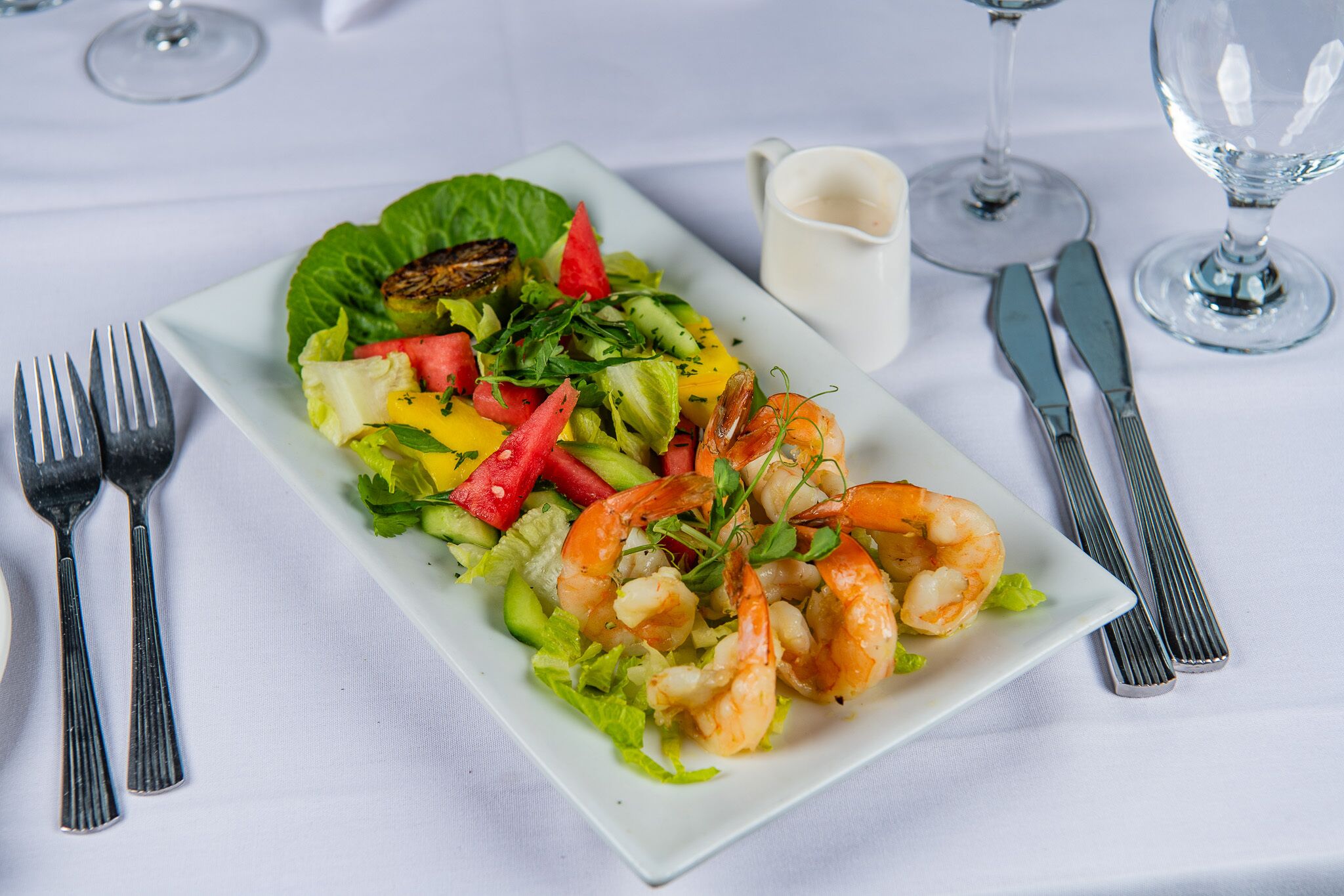 A rectangular plate with a salad and shrimp, accompanied by cutlery and a glass of white wine on a tablecloth.