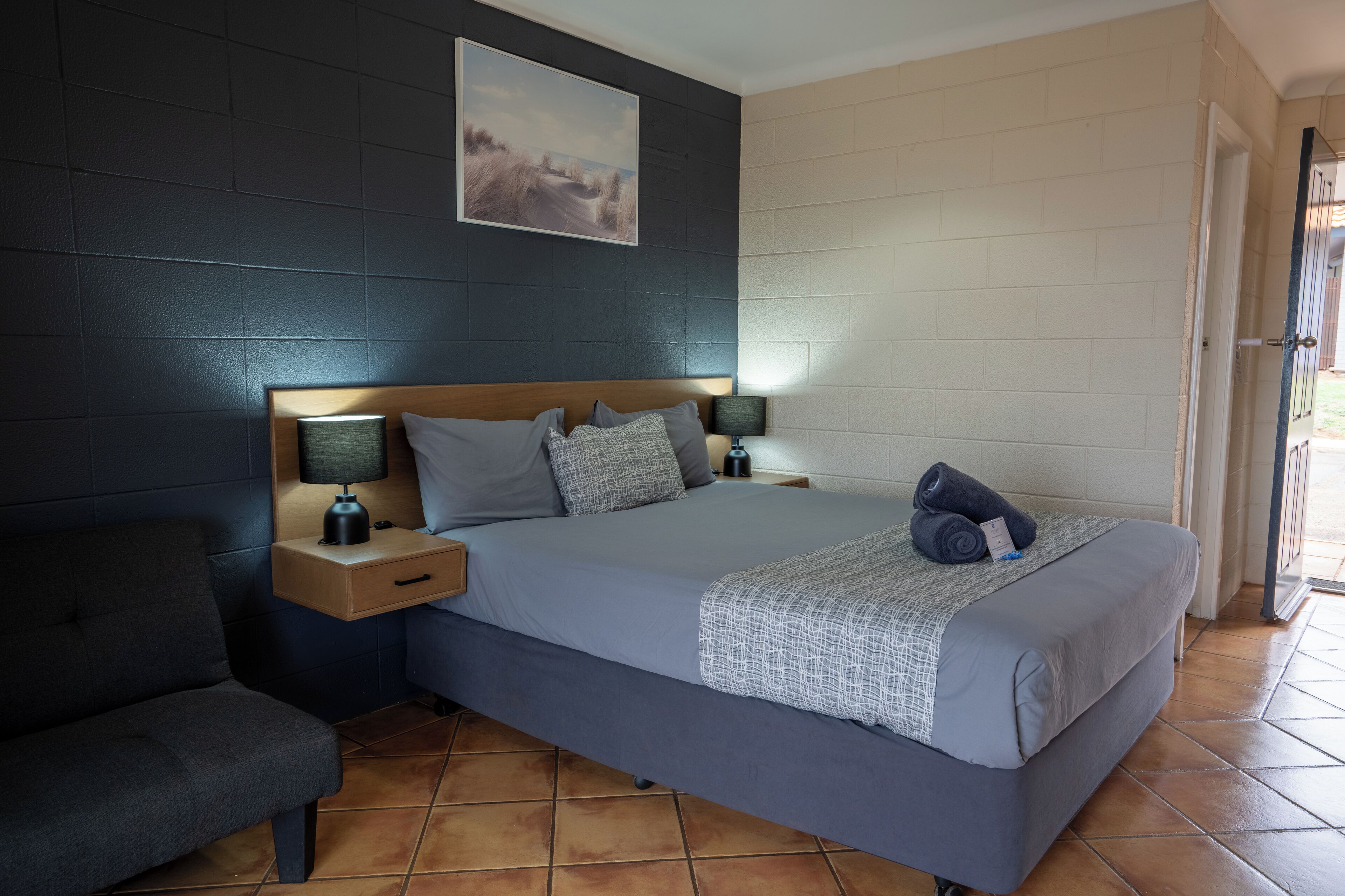 A neatly arranged bedroom with a bed, two bedside tables with lamps, a chair, and a painting on the wall.
