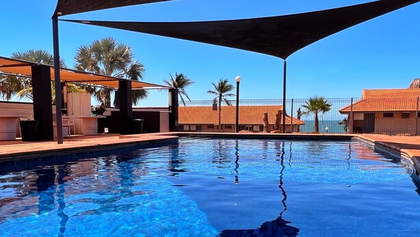 A clear blue pool with tiled flooring, surrounded by palm trees and a shaded area with outdoor furniture. - Hospitality Port Hedland (Port Hedland)