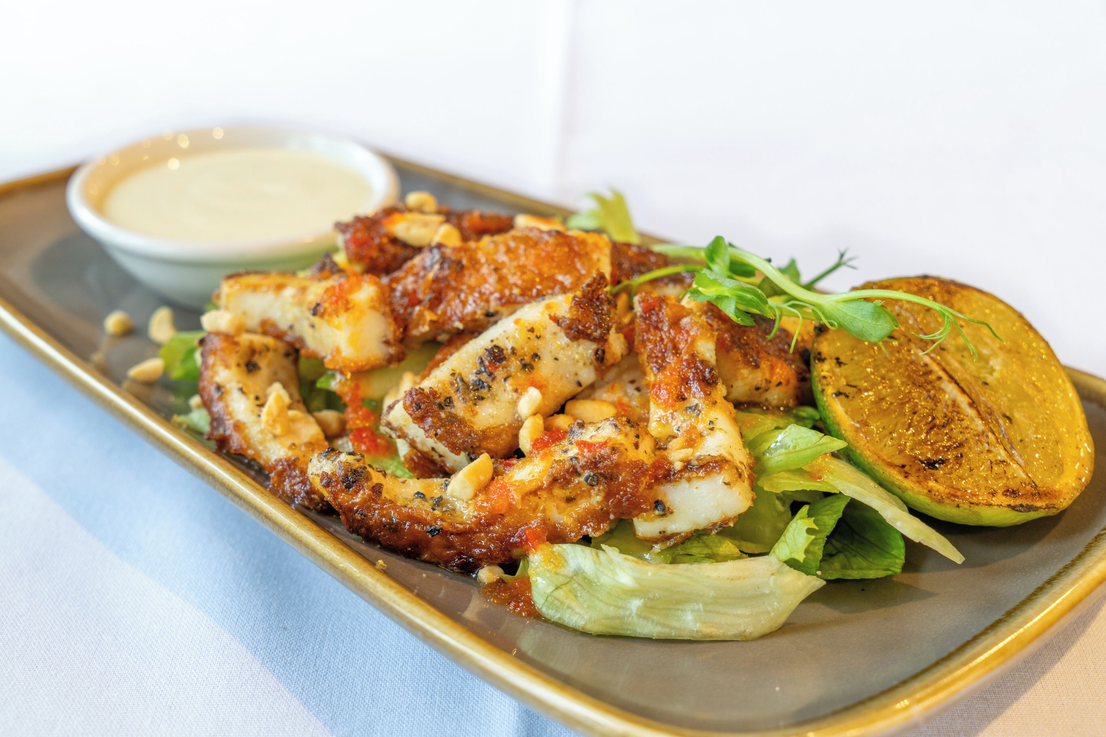 A plate of grilled chicken with a side of greens and a lemon wedge, served on a rectangular plate with a small bowl of sauce.