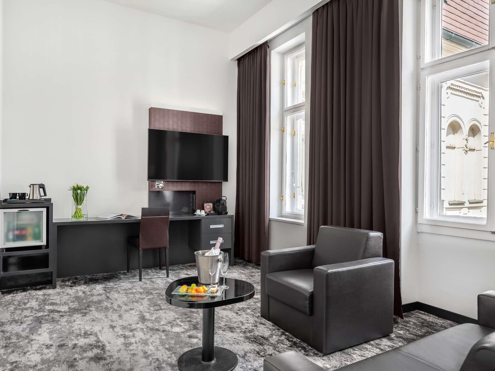 Une chambre d’hôtel moderne équipée d’un téléviseur à écran plat, d’un bureau avec une chaise, d’un canapé, d’une petite table ronde avec des boissons et des fruits, et de grandes fenêtres avec des rideaux.