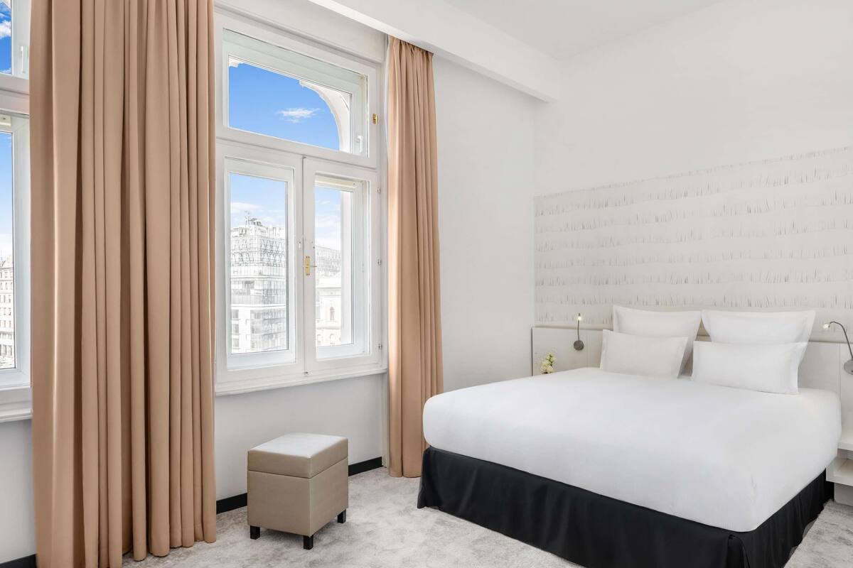 A hotel room with a large bed, a small ottoman, a nightstand, and a window with curtains.