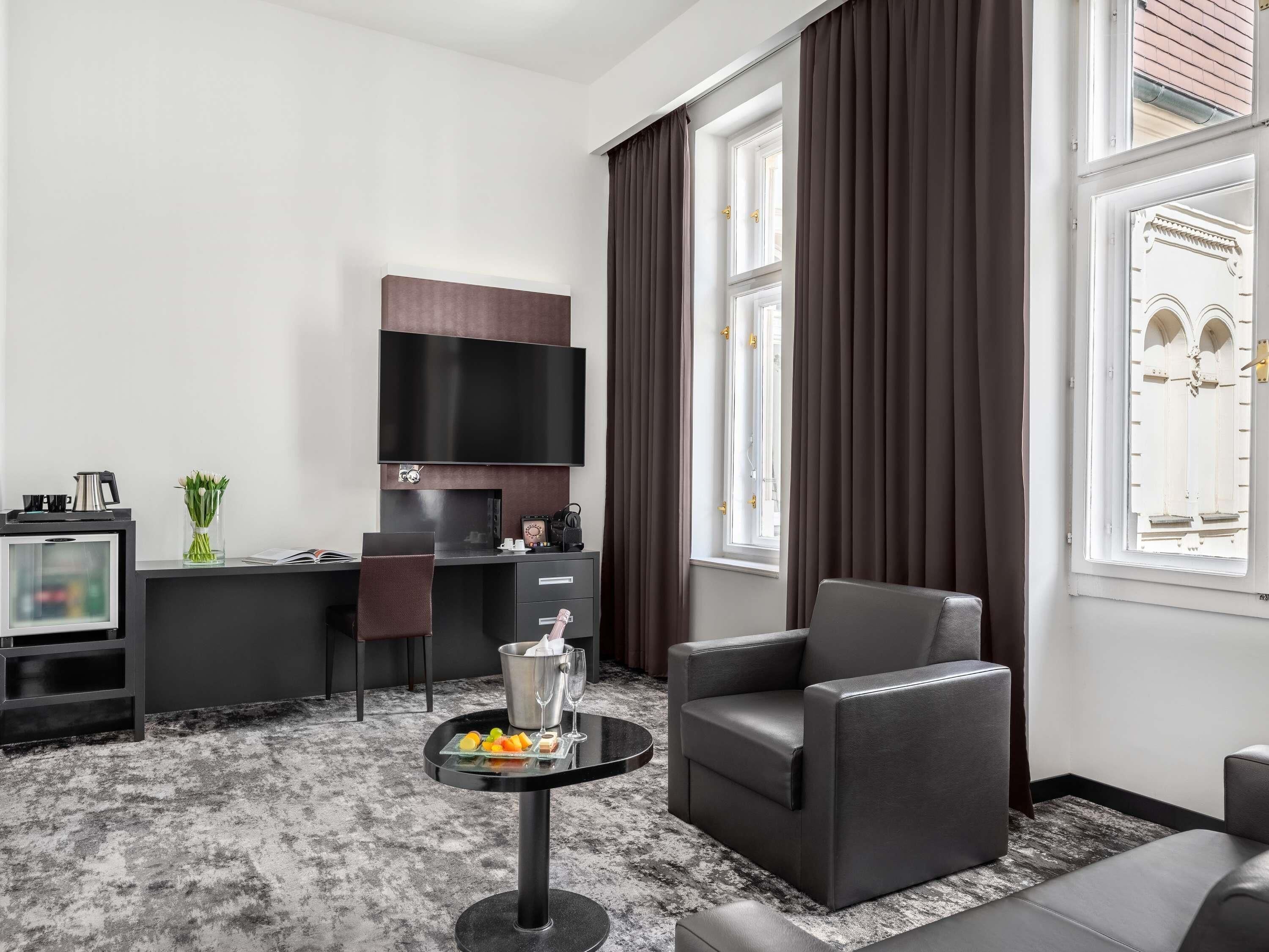 Une chambre d’hôtel moderne équipée d’un téléviseur à écran plat, d’un bureau avec une chaise, d’un canapé, d’une petite table ronde avec des boissons et des en-cas, et de grandes fenêtres avec des rideaux.
