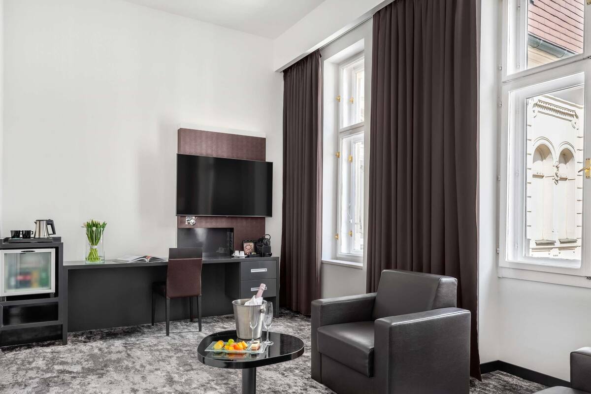 A modern hotel room with a flat-screen TV, a desk with a chair, a sofa, a small round table with a drink and snacks, and large windows with curtains.