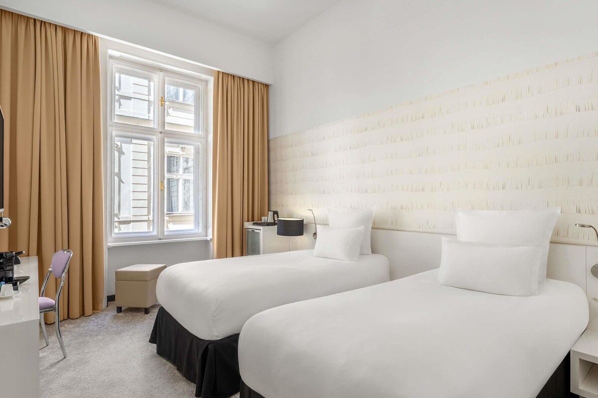 Une chambre d’hôtel avec deux lits, un bureau, une chaise, une lampe et une fenêtre avec des rideaux.
