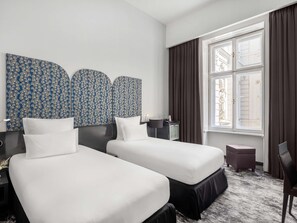 Habitación de hotel con dos camas, un ventanal con cortinas, una mesita de noche con lámpara y un panel decorativo en la pared.