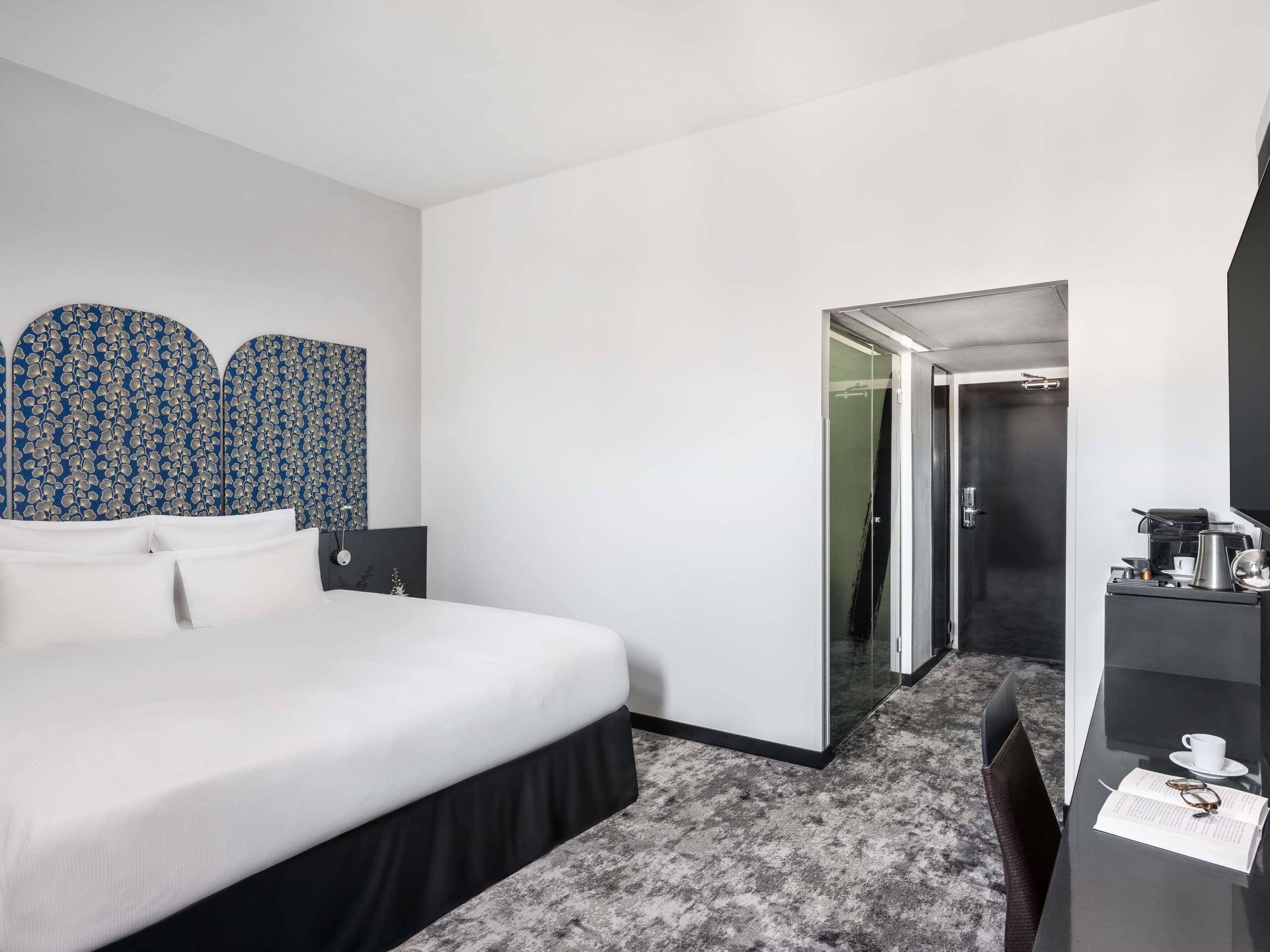 Quarto de hotel com cama, escrivaninha, cadeira, cafeteira e porta para um banheiro.