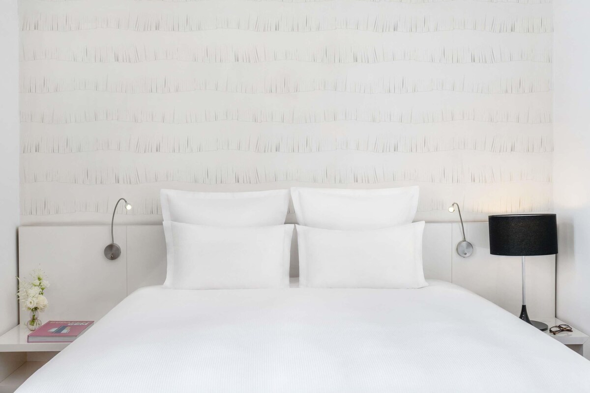 Un lit bien fait, avec du linge de lit blanc, une table de chevet avec une lampe et un mur orné d’un motif texturé.