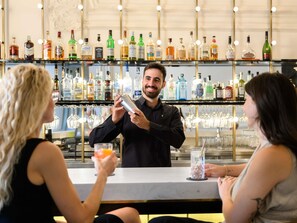 Zona de bar con un barman preparando una bebida, estantes con diferentes botellas de licor y copas de vino colgando.