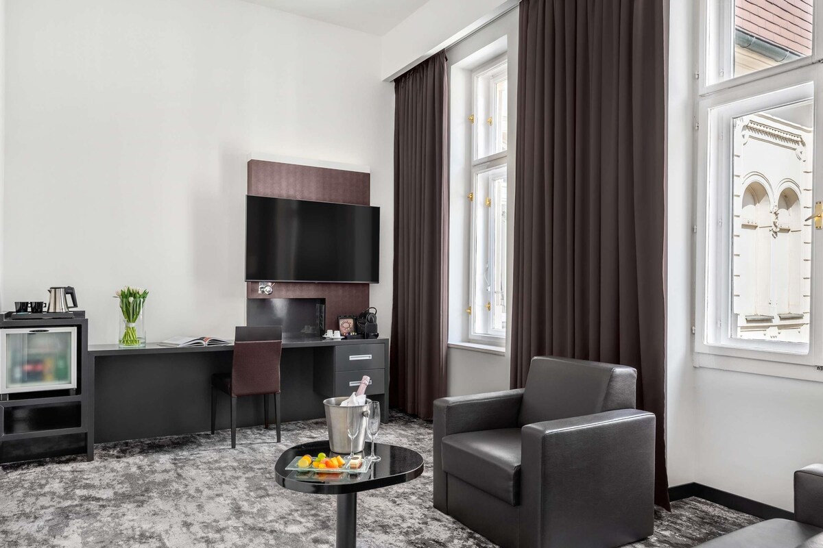 Une chambre d’hôtel moderne équipée d’un téléviseur à écran plat, d’un bureau avec une chaise, d’un canapé, d’une petite table ronde avec des boissons et des en-cas, et de grandes fenêtres avec des rideaux.