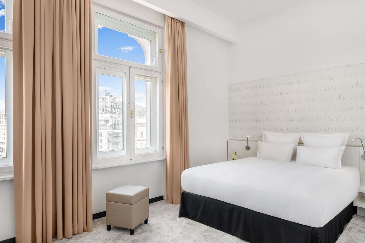 Une chambre d’hôtel avec un grand lit, un petit pouf, une table de chevet et une fenêtre avec des rideaux.