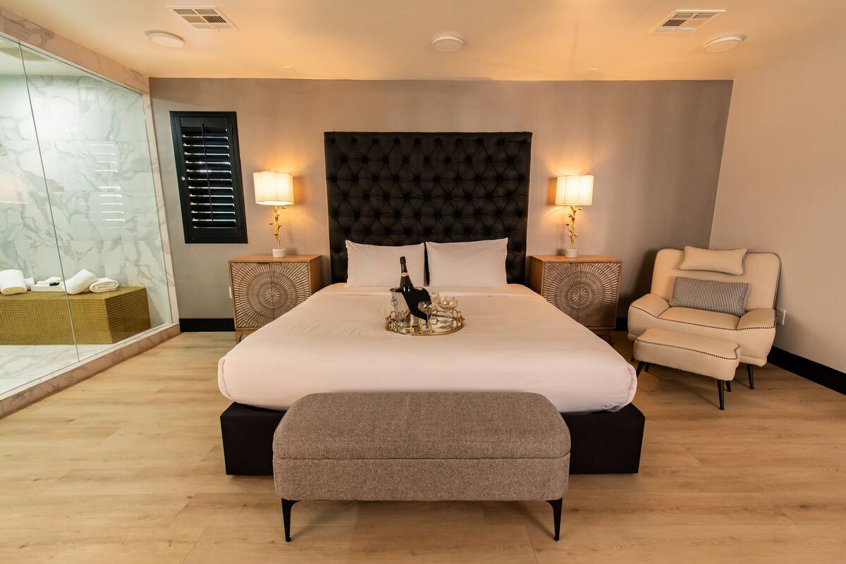 A neatly arranged bedroom with a large bed, bedside lamps, a chair, and a glass partition.