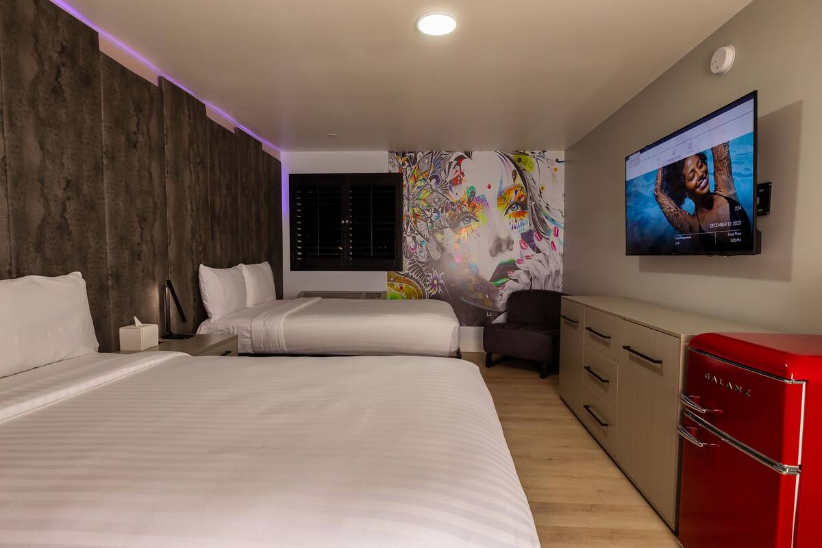 A modern hotel room with a large bed, a desk, a chair, a TV, and a red minibar.