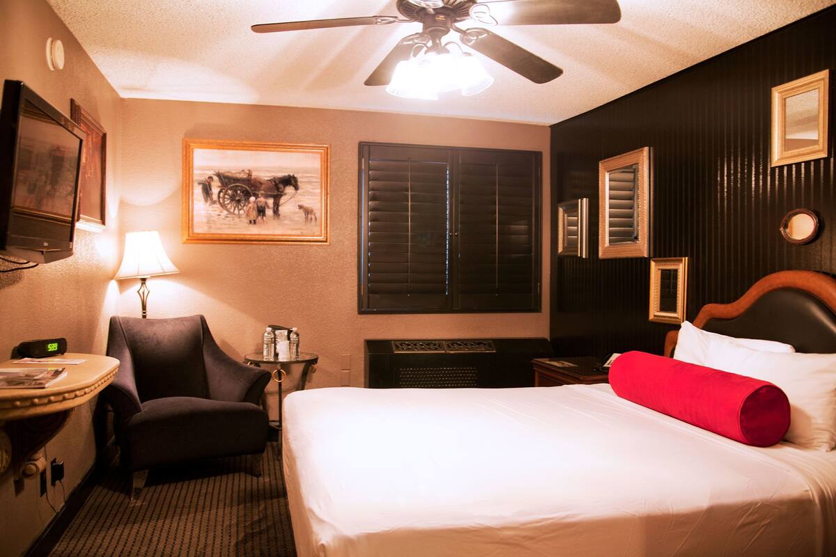 A hotel room with a bed, a chair, a desk, a lamp, a ceiling fan, and a framed picture on the wall.
