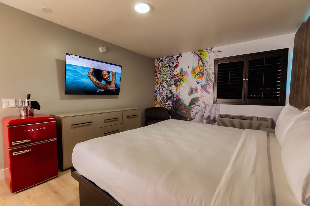 A hotel room with a bed, a red minibar, a television on the wall, and a window with blinds.