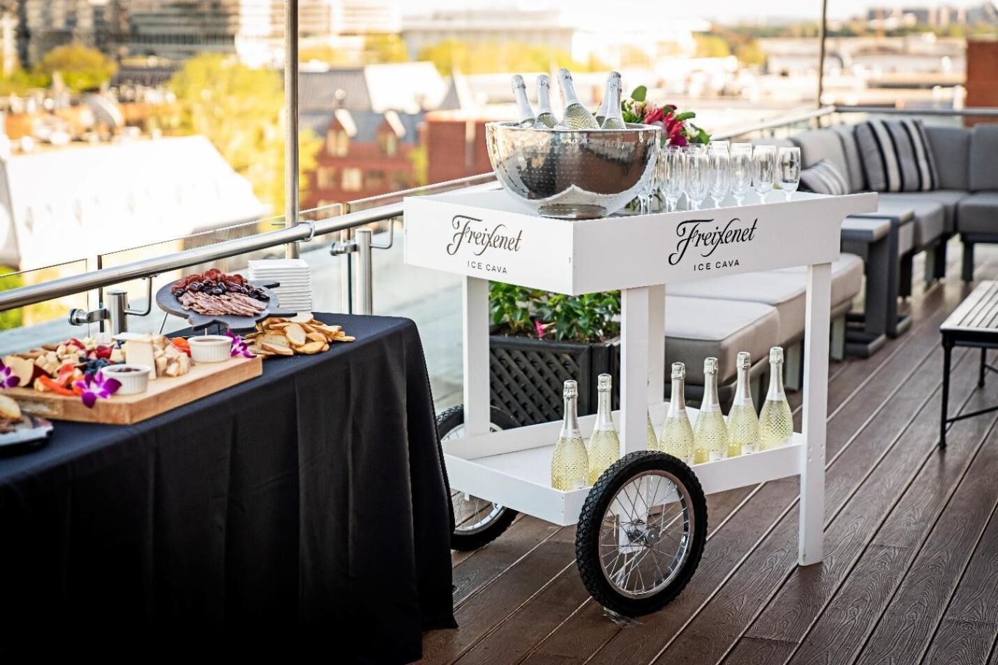 An outdoor event setup with a table for food and drinks, a cart labeled "Freixenet Ice Cava," and a city view in the background.