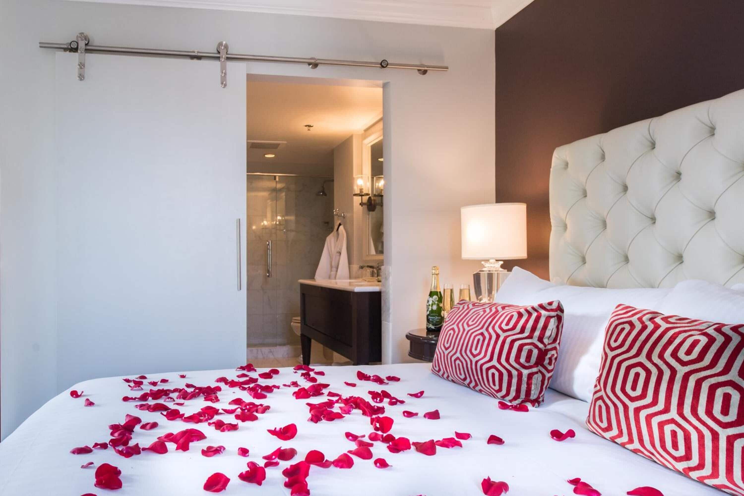 A hotel room with a bed covered in rose petals, a tufted headboard, and a nightstand with a lamp and champagne bottle.