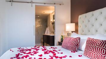A hotel room with a bed covered in rose petals, a tufted headboard, and a nightstand with a lamp and champagne bottle.