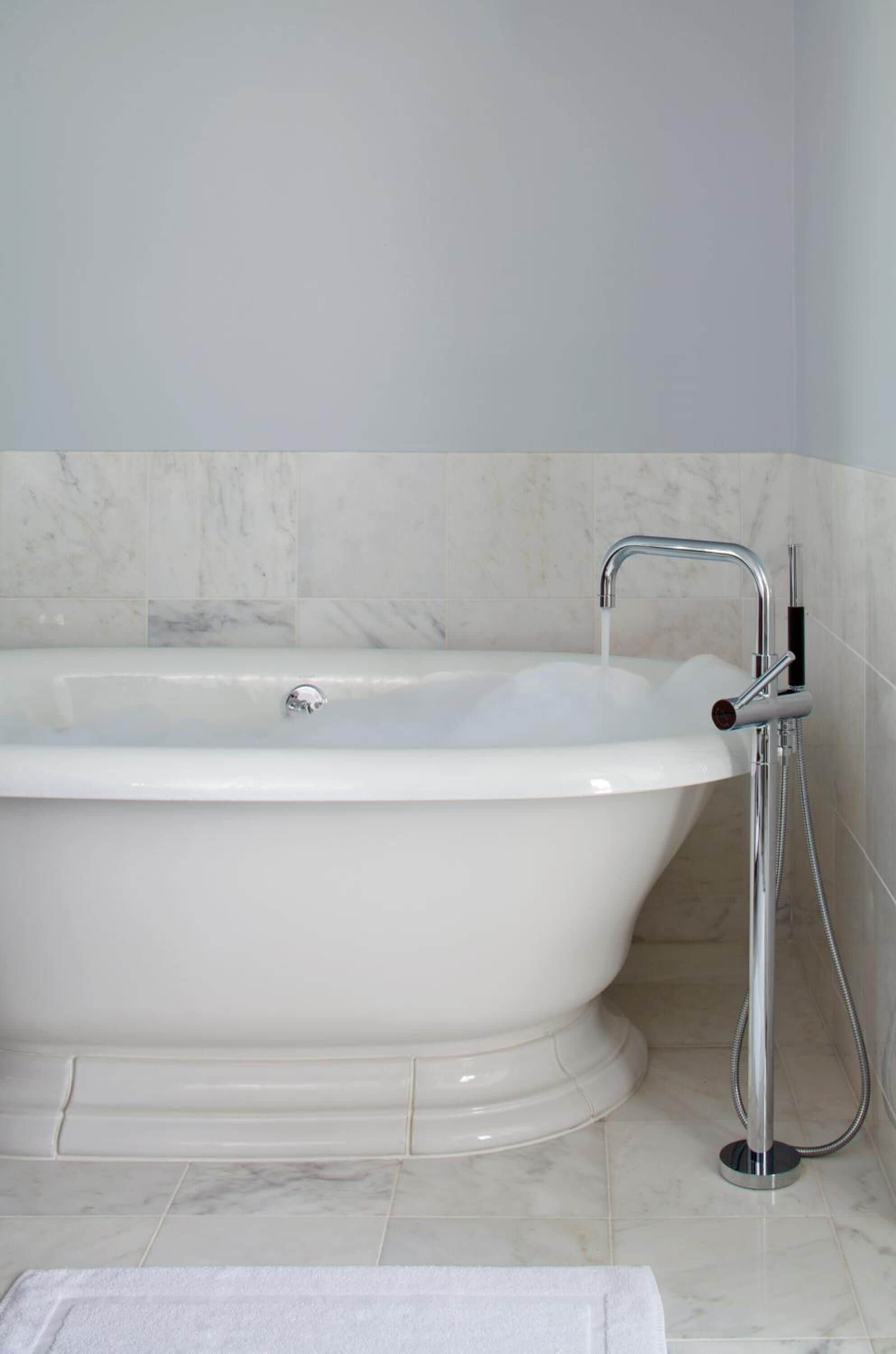 A white freestanding bathtub with a handheld showerhead, marble tiles, and a white towel on the floor.