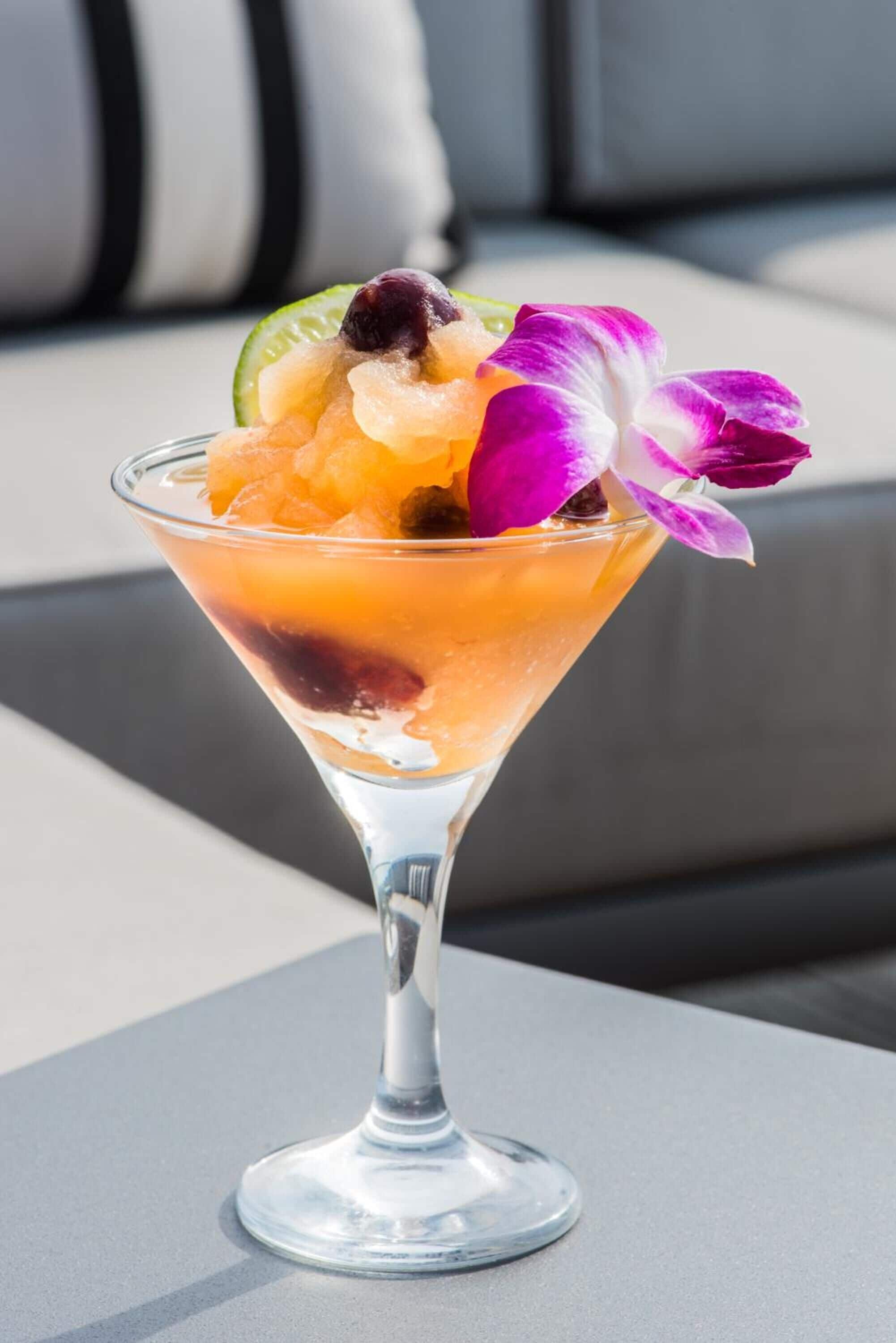 Un cóctel en una copa de martini con frutas y una flor morada, sobre una mesa con superficie gris.