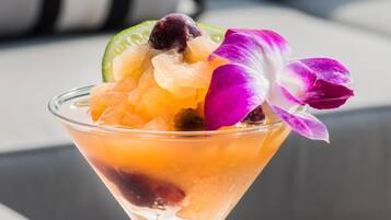 Un cóctel en una copa de martini con frutas y una flor morada, sobre una mesa con superficie gris.