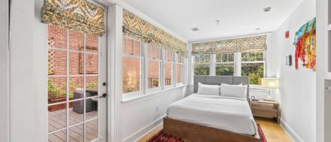 A bedroom with a large bed, a wooden floor, a red patterned rug, and a view of a brick wall and greenery outside.