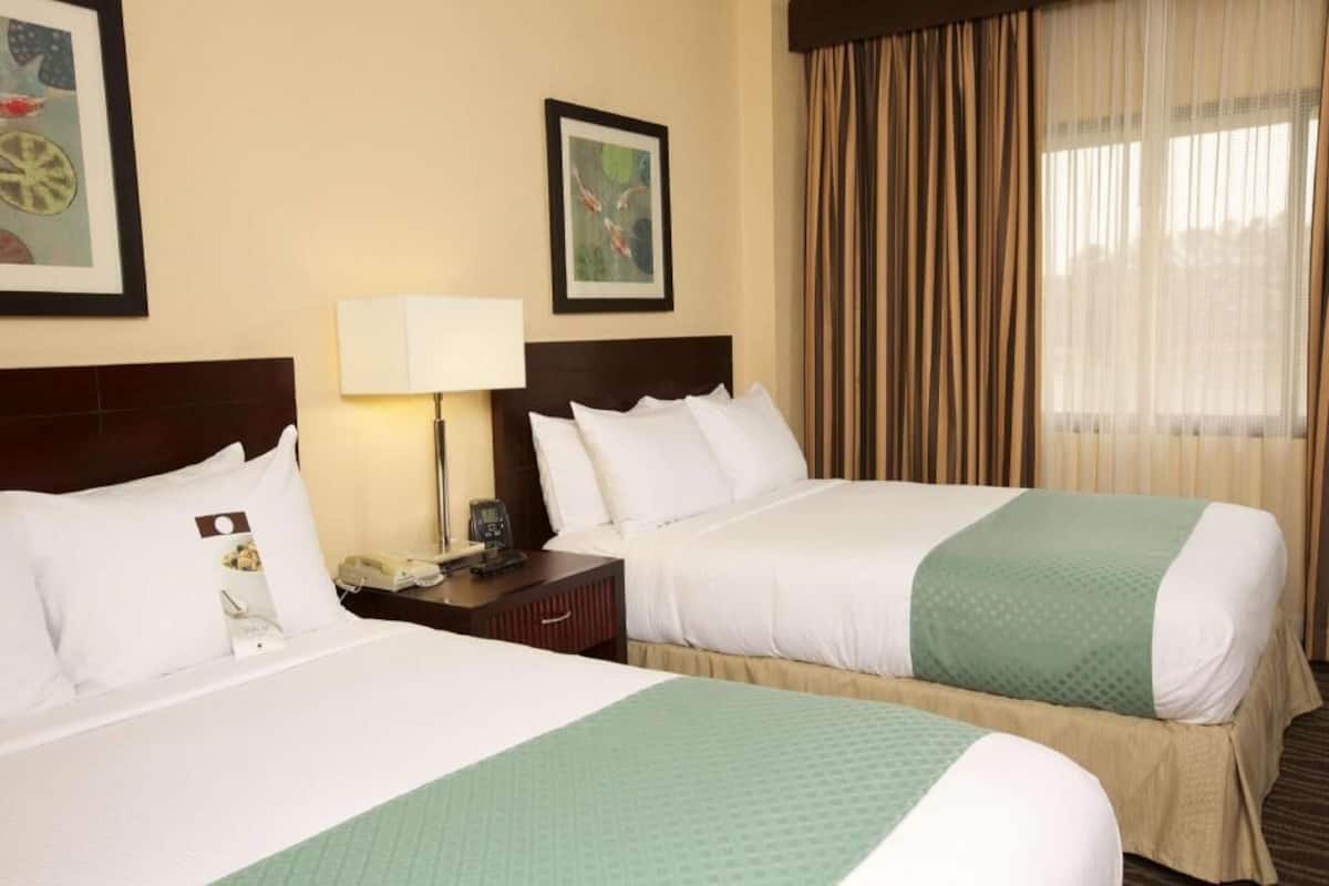 A hotel room with two beds, a nightstand with a lamp, and a window with curtains.