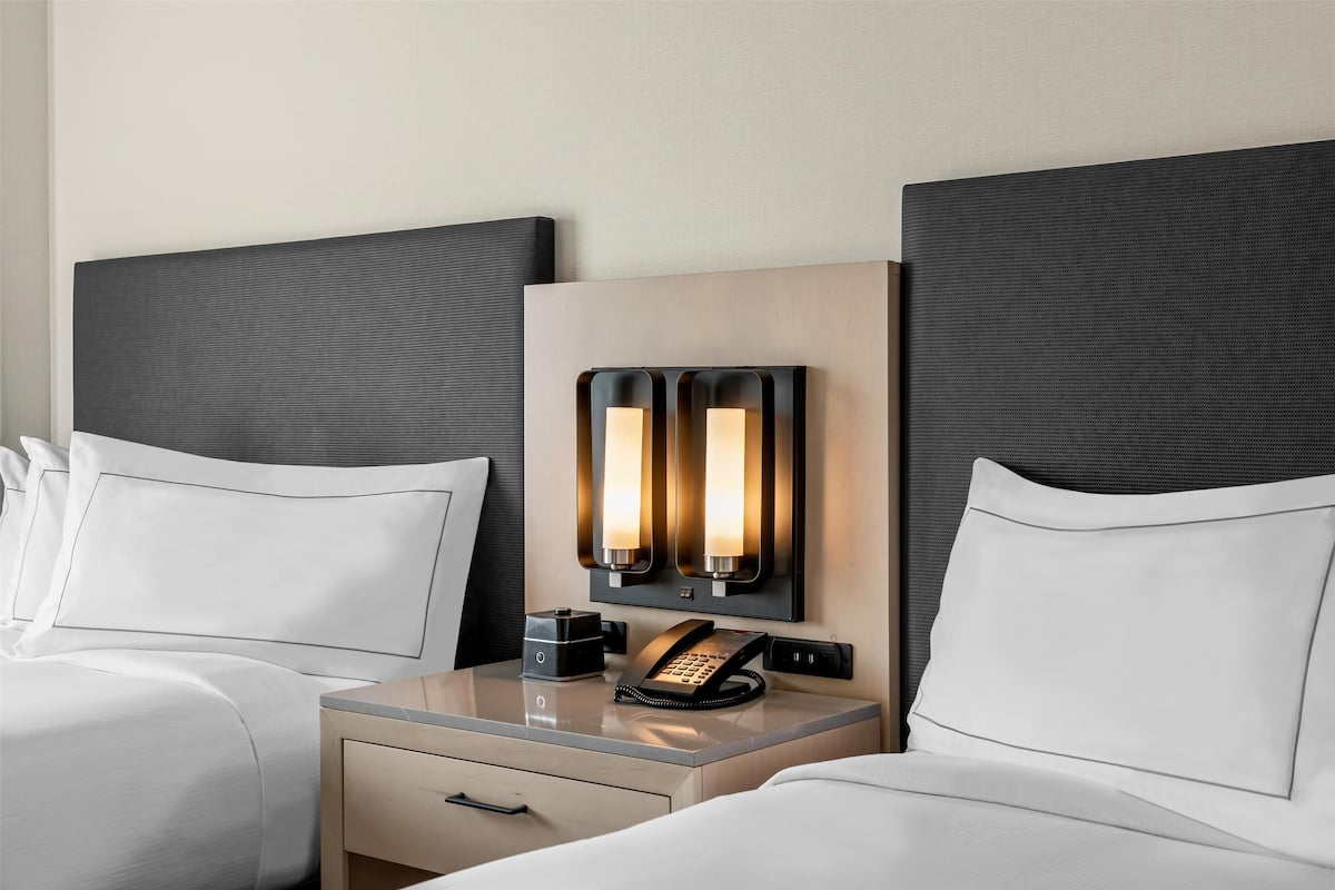 A hotel room with two beds, a bedside table with a phone and a coffee maker, and a wall-mounted light fixture.