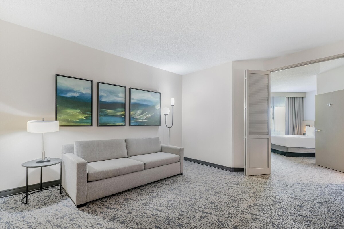 A hotel room with a grey sofa, a small table with a lamp, and three framed abstract paintings on the wall.