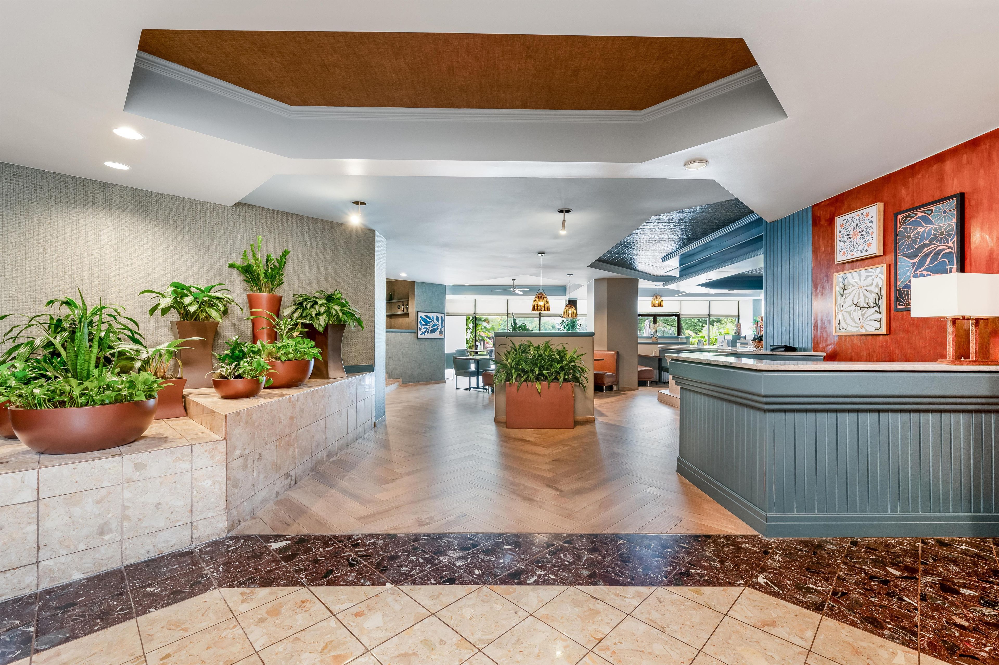 A hotel lobby with a reception desk, potted plants, a herringbone floor pattern, and a view of the dining area.