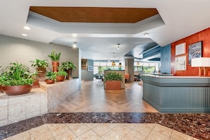 A hotel lobby with a reception desk, potted plants, a herringbone floor pattern, and a view of the dining area. - DoubleTree Suites by Hilton Raleigh - Durham (Durham)