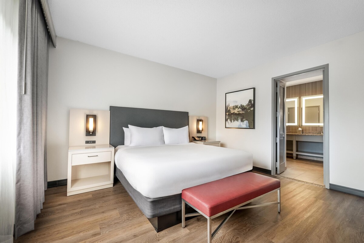A hotel room with a bed, bedside tables, a bench, a painting, and a bathroom visible through an open door.