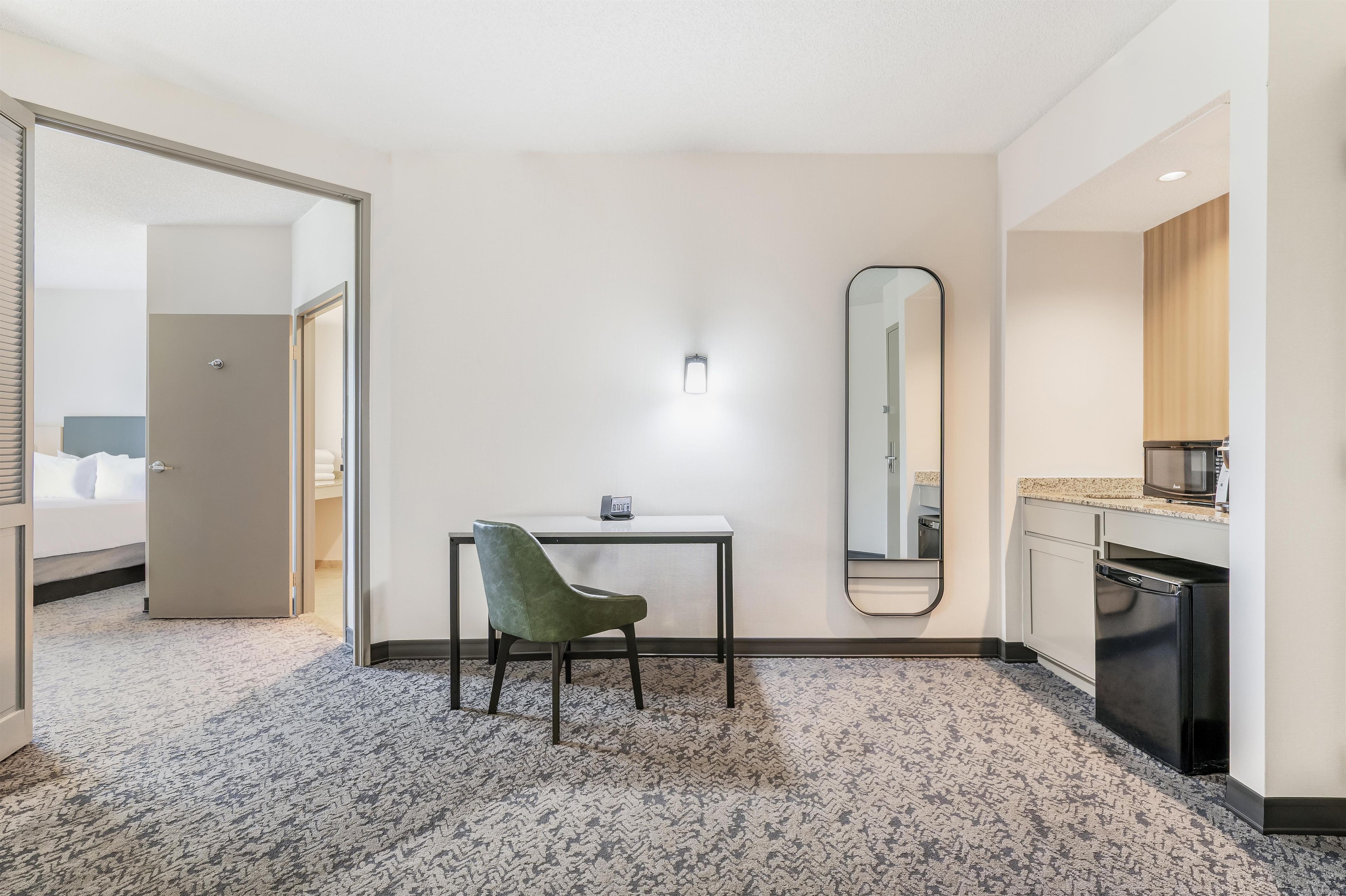 Ein Hotelzimmer mit einem Bett, einem Schreibtisch mit Stuhl, einem Spiegel, einer kleinen Küchenzeile und einem durch eine geöffnete Tür einsehbaren Badezimmer.