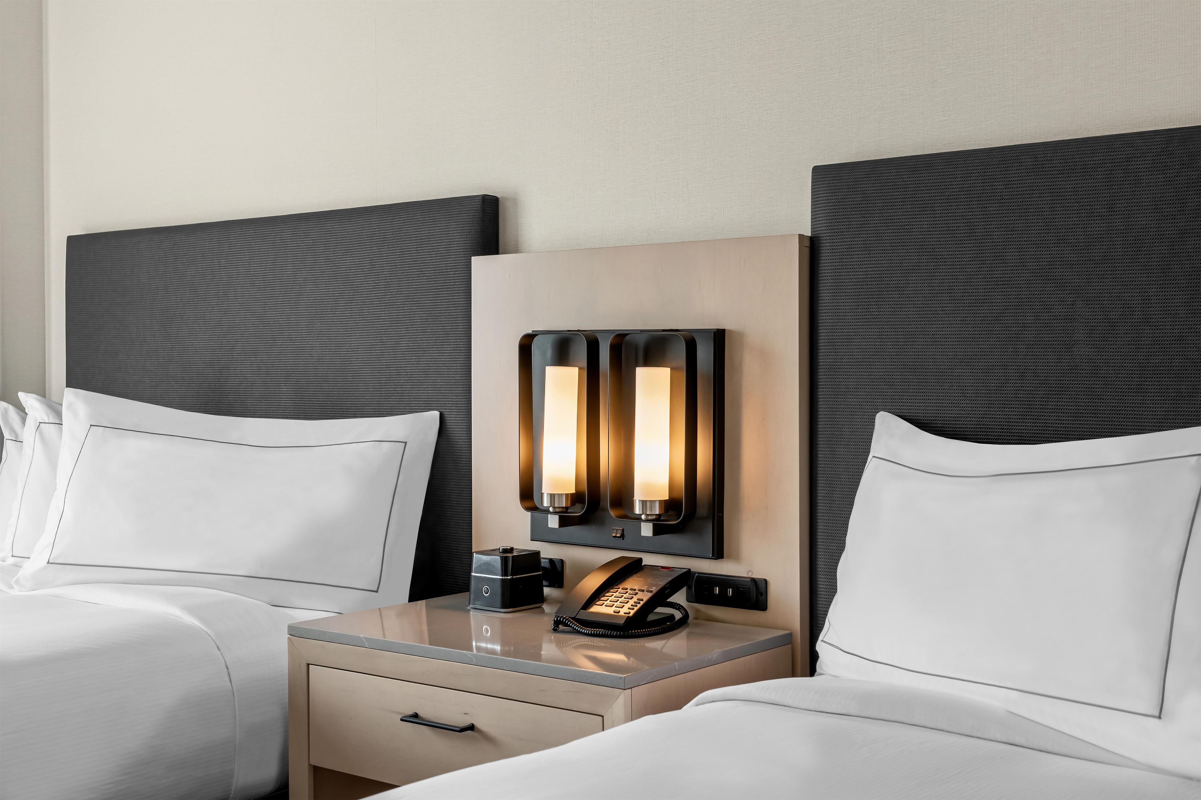A hotel room with two beds, a bedside table with a lamp, a telephone, and a small drawer.