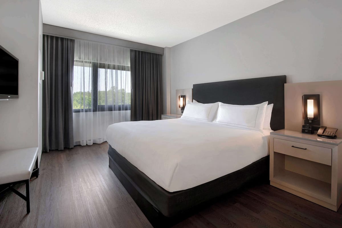 A hotel room with a large bed, a bedside table with a lamp, a TV mounted on the wall, and a window with curtains.