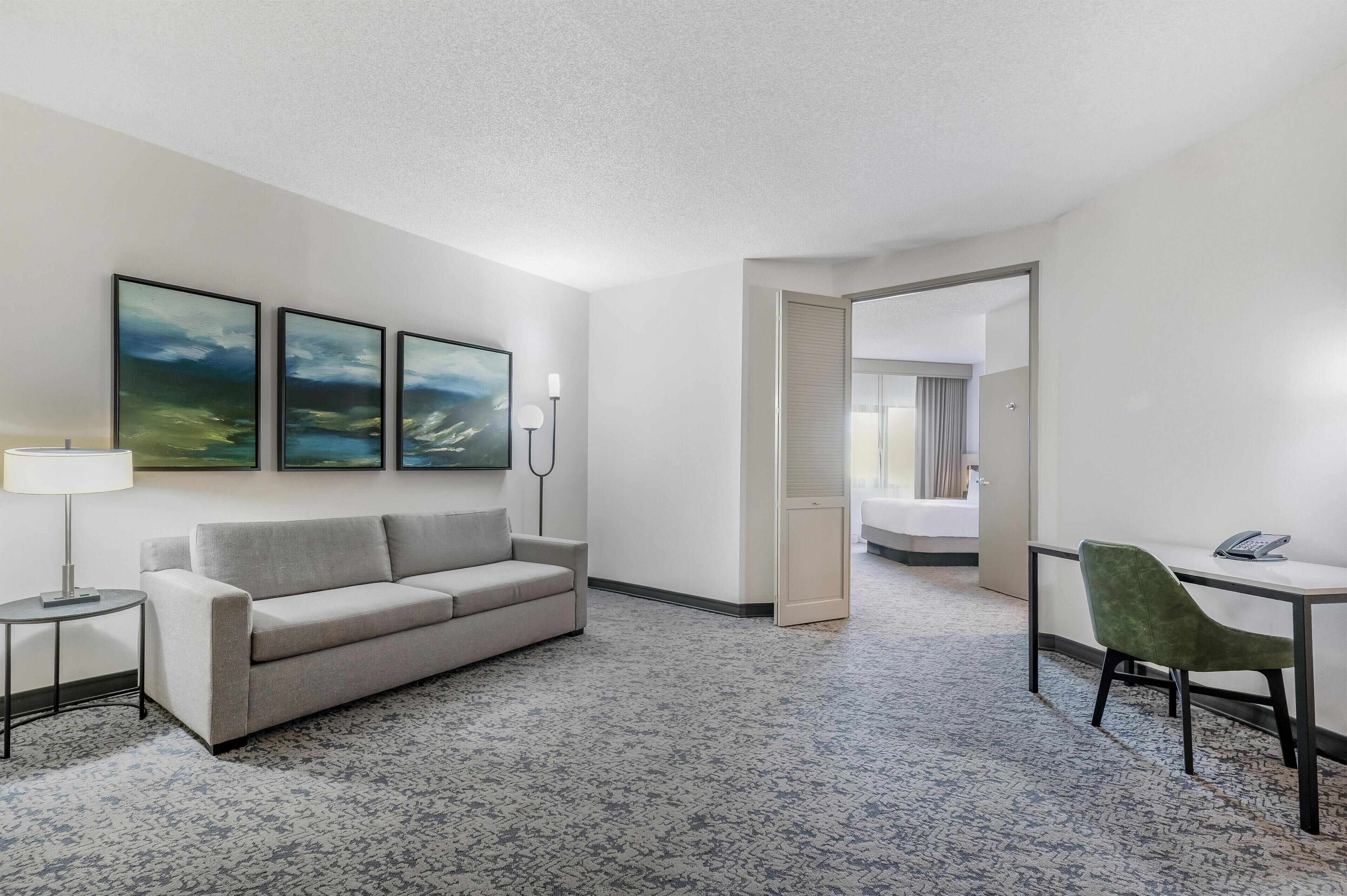 A hotel room with a gray sofa, a small table with a lamp, a desk with a chair, and a bed in the background.