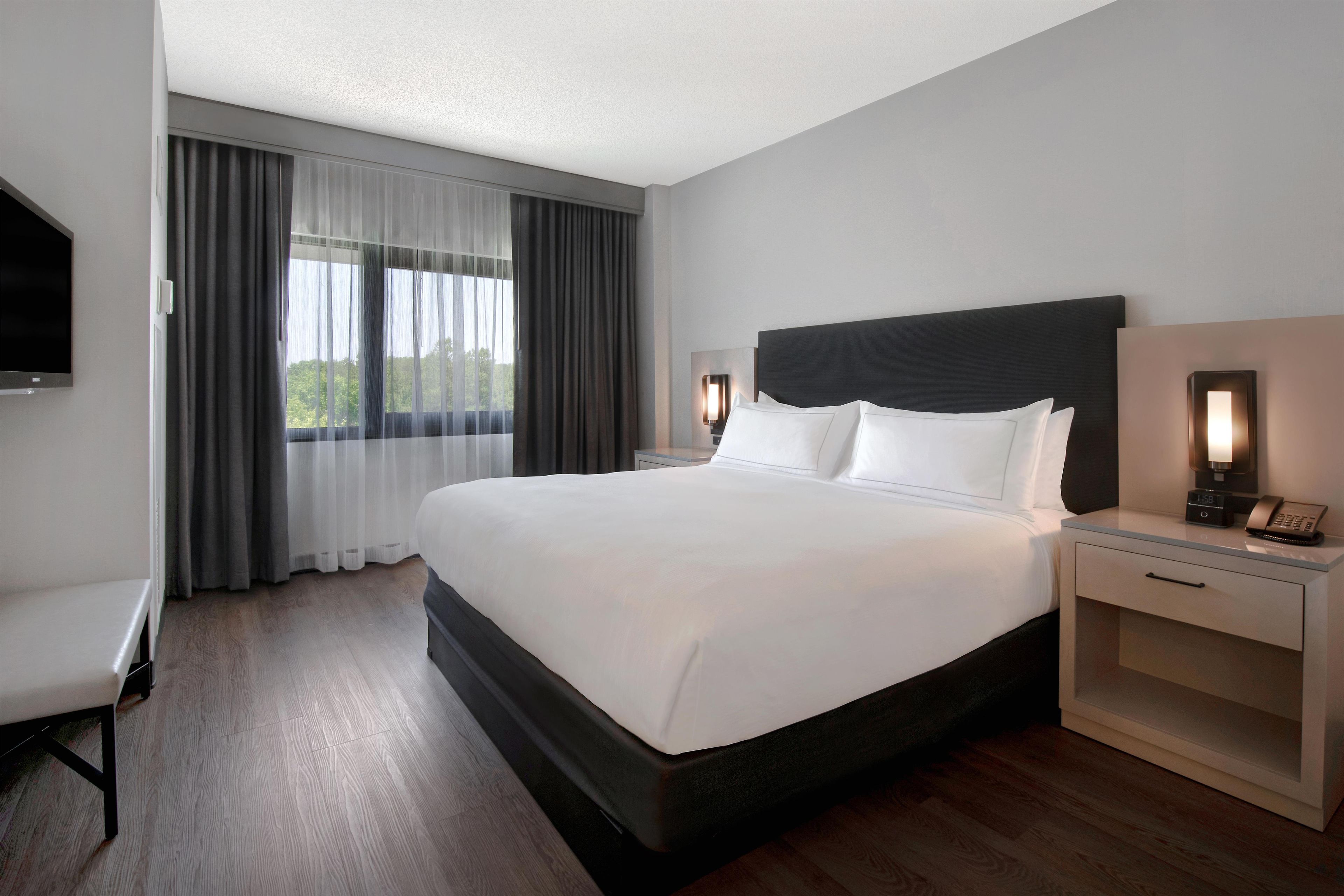 Habitación de hotel con una cama grande, una mesita de noche con lámpara, televisor y ventana con cortinas.