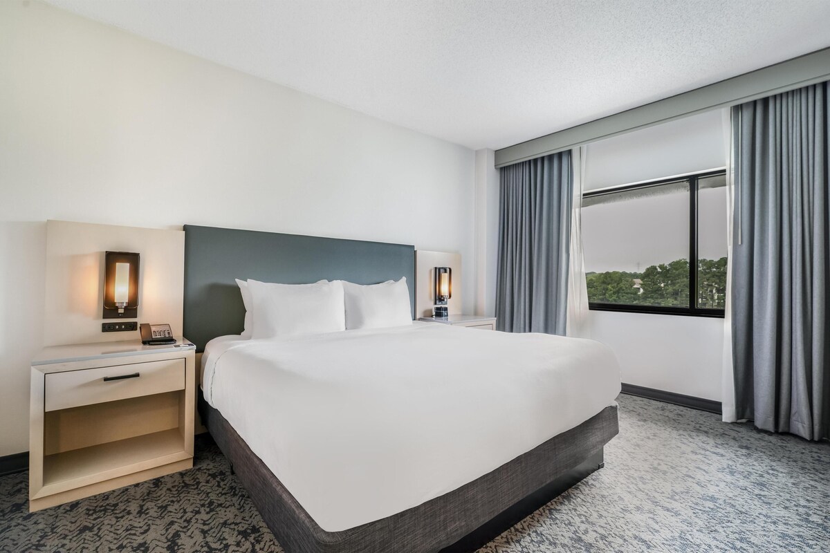 A hotel room with a large bed, two bedside tables with lamps, a window with curtains, and a view of the outdoors.