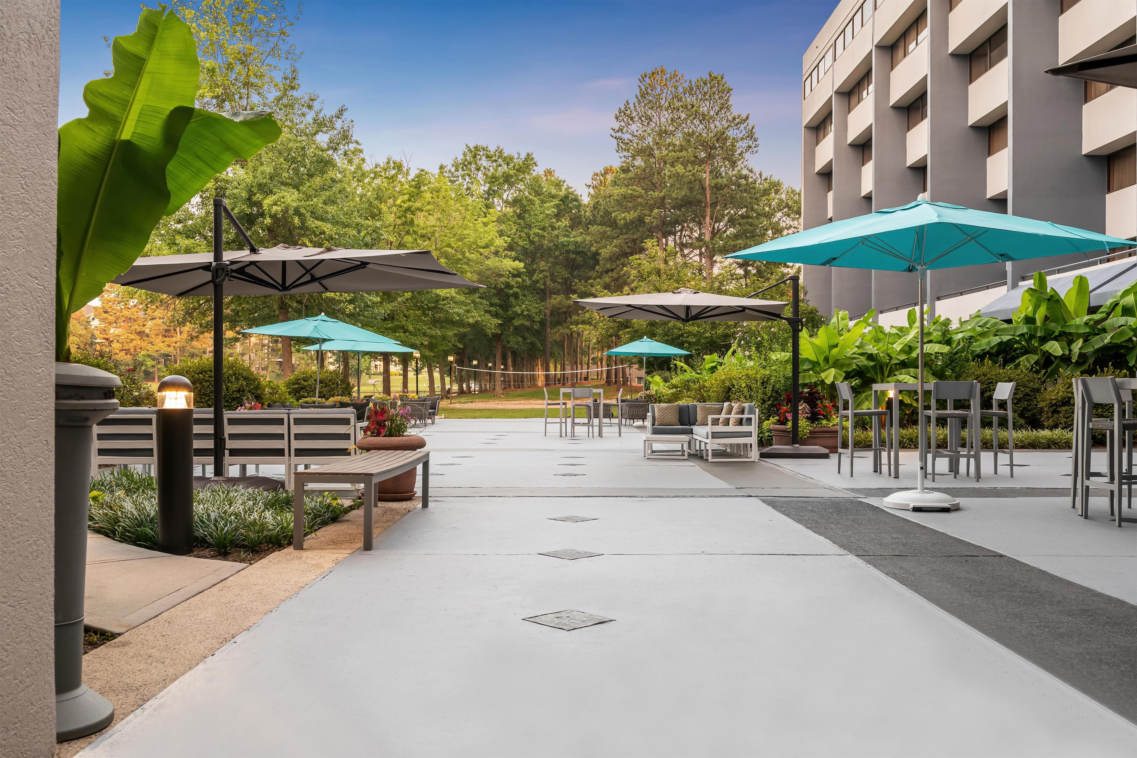 An outdoor patio area with seating and umbrellas, surrounded by greenery and a building.