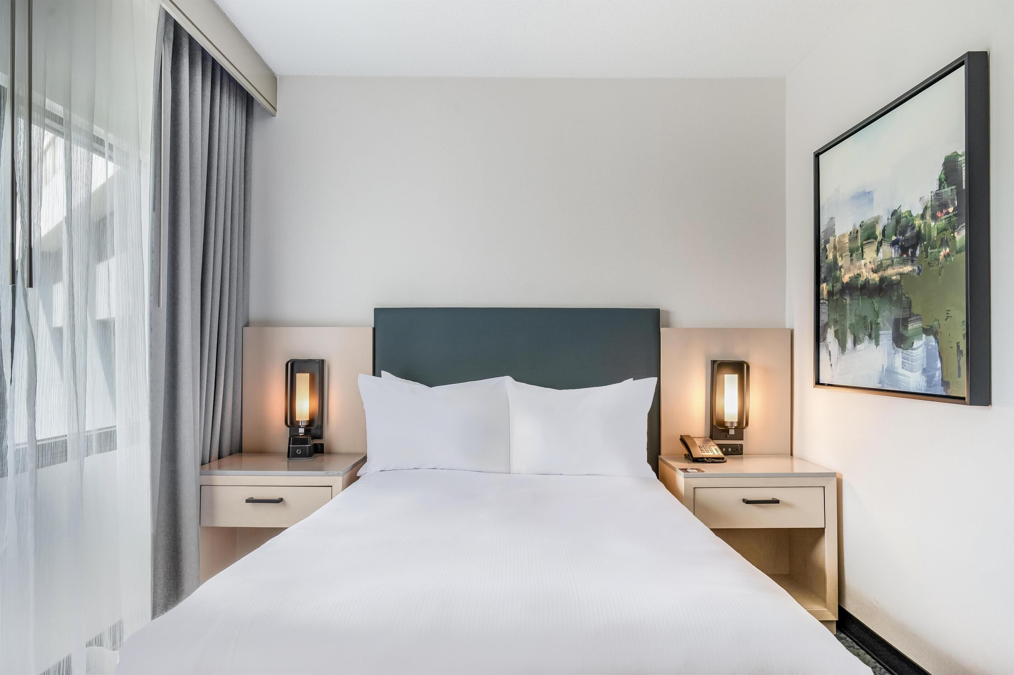 A hotel room with a large bed, two bedside tables with lamps, a window with curtains, and a painting on the wall.