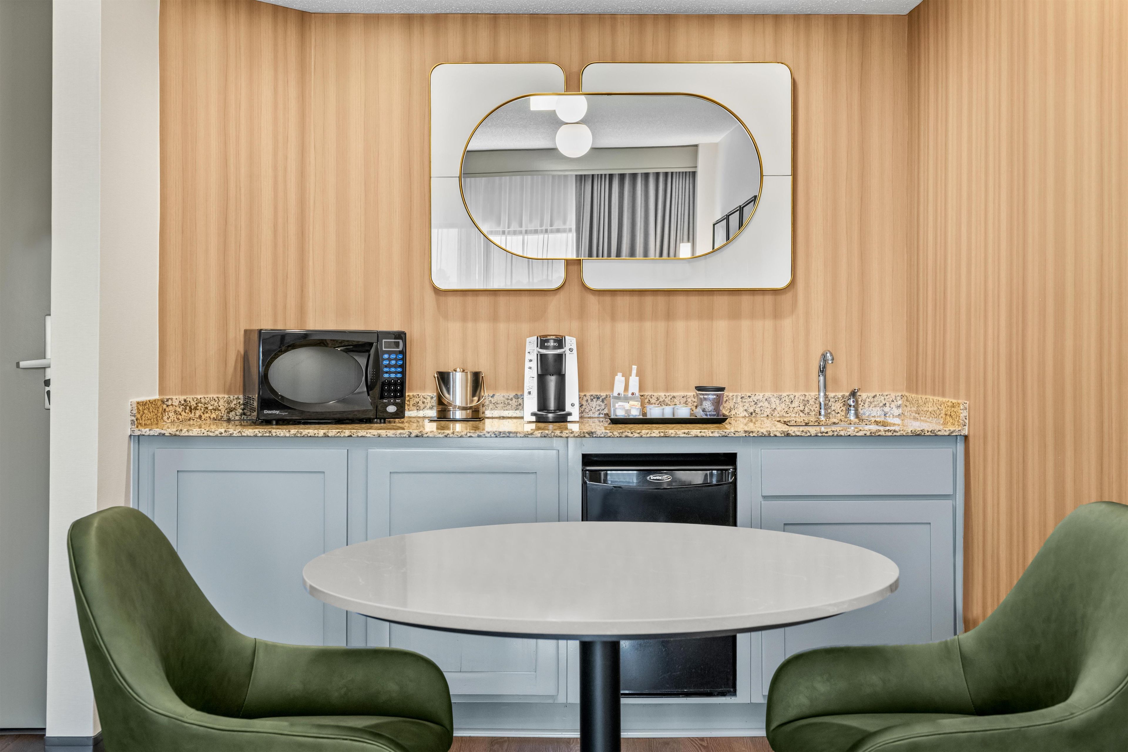 Cozinha compacta com micro-ondas, cafeteira e máquina de lavar loiça, uma mesa redonda com duas cadeiras e um espelho sobre o balcão.