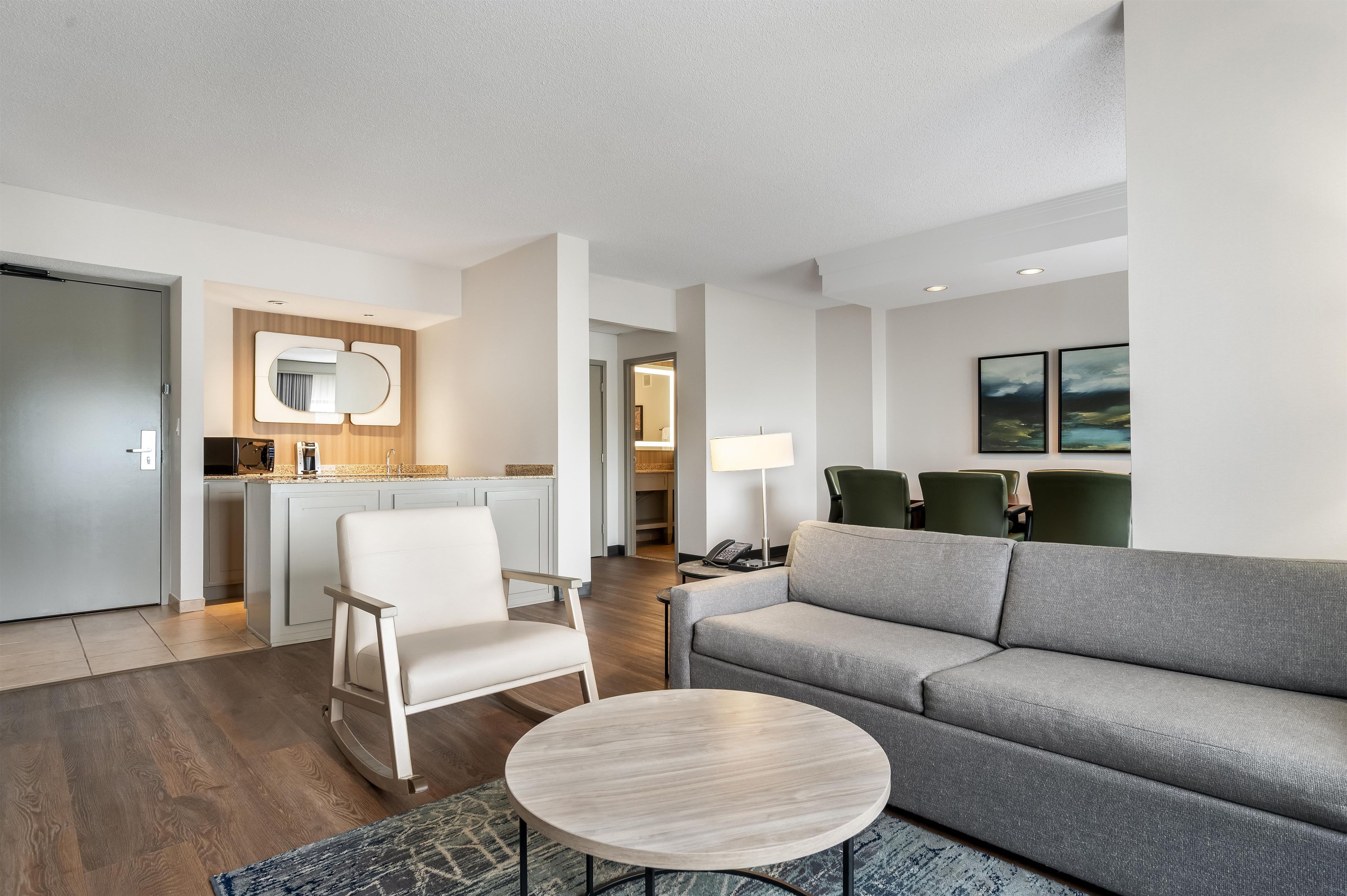 Quarto de hotel moderno com sofá, uma cadeira, uma mesinha, cozinha e área de estar com cadeiras.