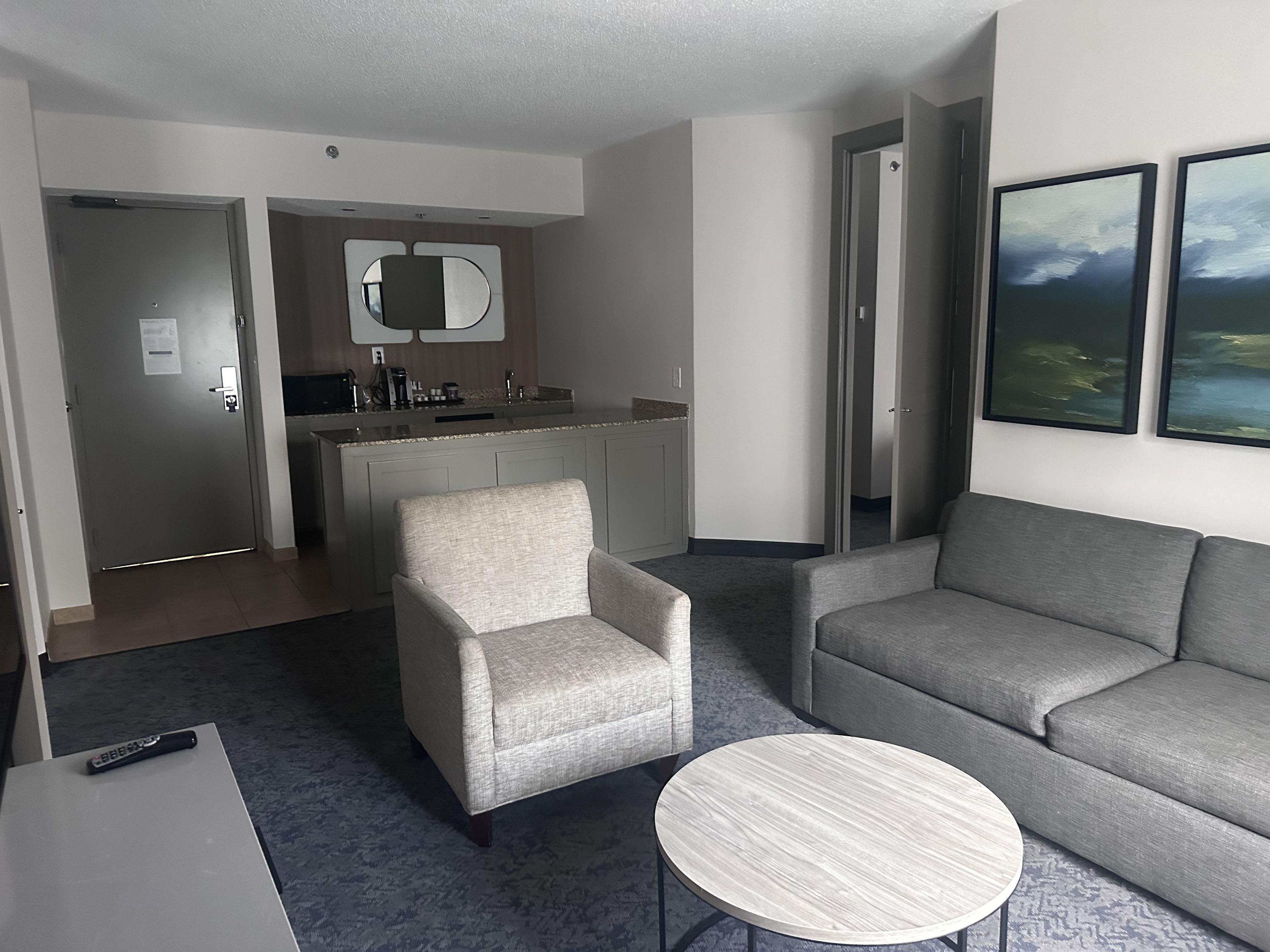 Ein Hotelzimmer mit grauem Sofa, grauem Sessel, rundem Couchtisch, einer Küchenzeile mit Spiegel und Blick auf das Meer durch ein Fenster.